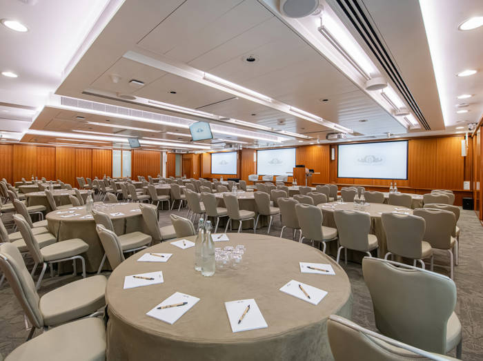 A well-lit conference room with round tables, chairs, notepads, and pens, featuring large screens and a podium for events.